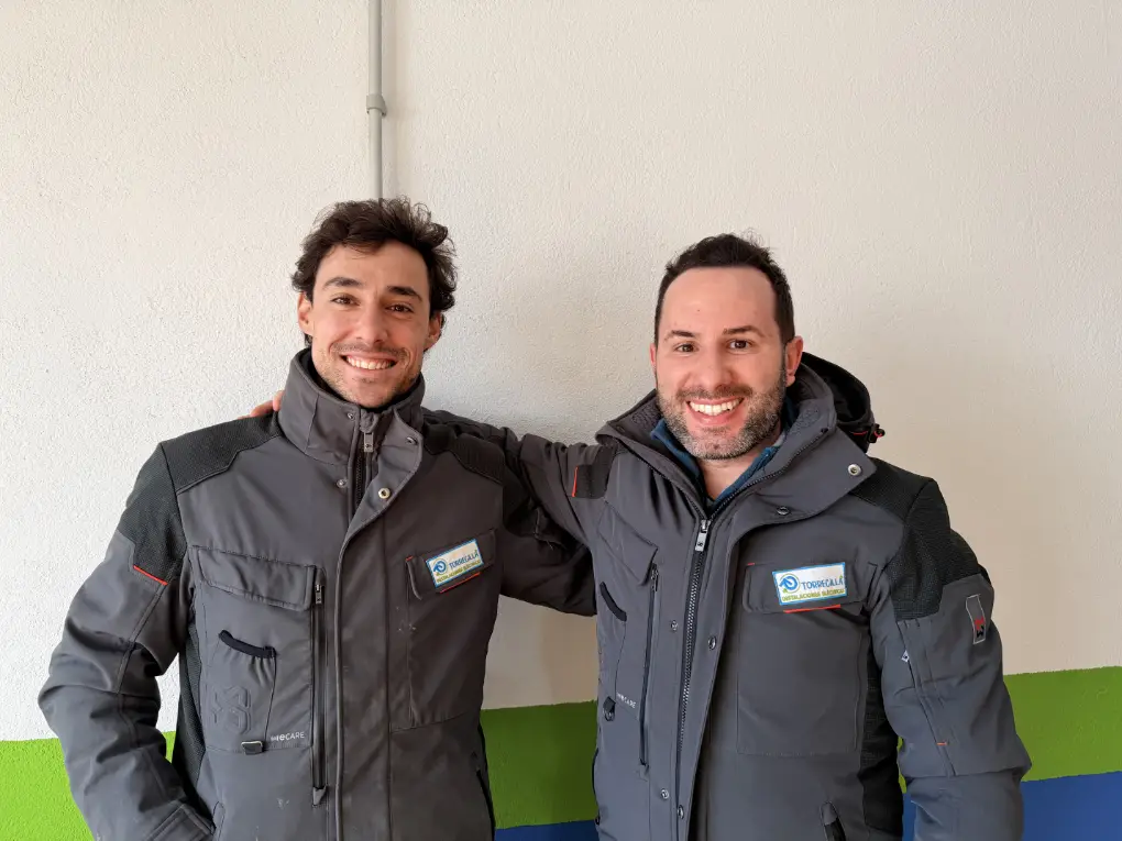 Plano medio de dos técnicos de servicios eléctricos uniformados y sonrientes, posando juntos en un entorno profesional.