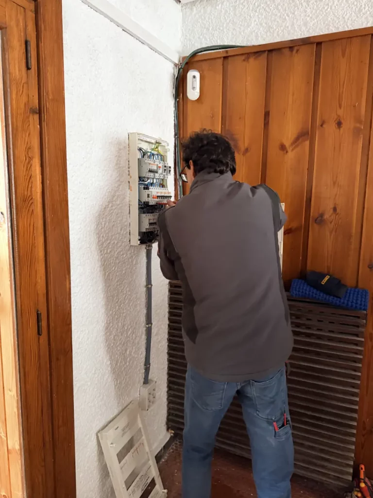 Un técnico electricista de espaldas trabajando en un cuadro eléctrico abierto en la pared de una vivienda. Se observa el cableado y los interruptores automáticos mientras realiza la instalación.