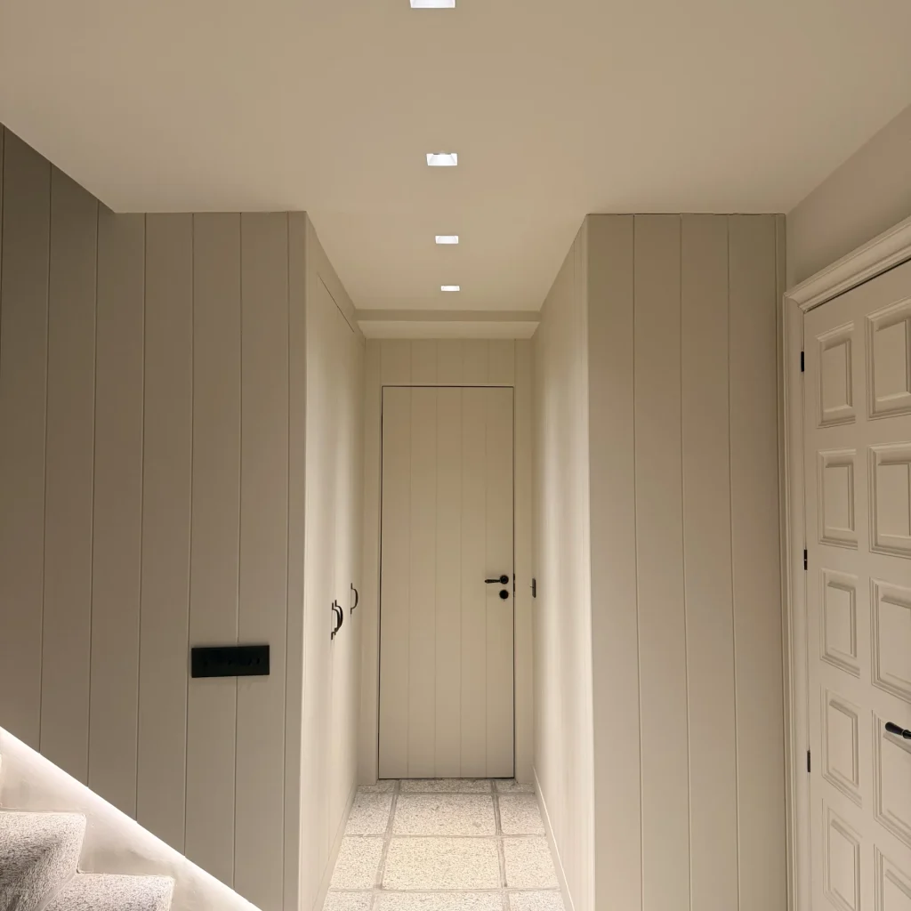 Vista más cercana del pasillo y las puertas. Las paredes están revestidas en paneles verticales de color crema. El techo tiene luces empotradas. Una puerta simple de color claro se ve al final del pasillo, y a la derecha, una puerta de entrada con paneles decorativos. El suelo es de baldosas claras. Se aprecia el comienzo de la escalera a la izquierda.