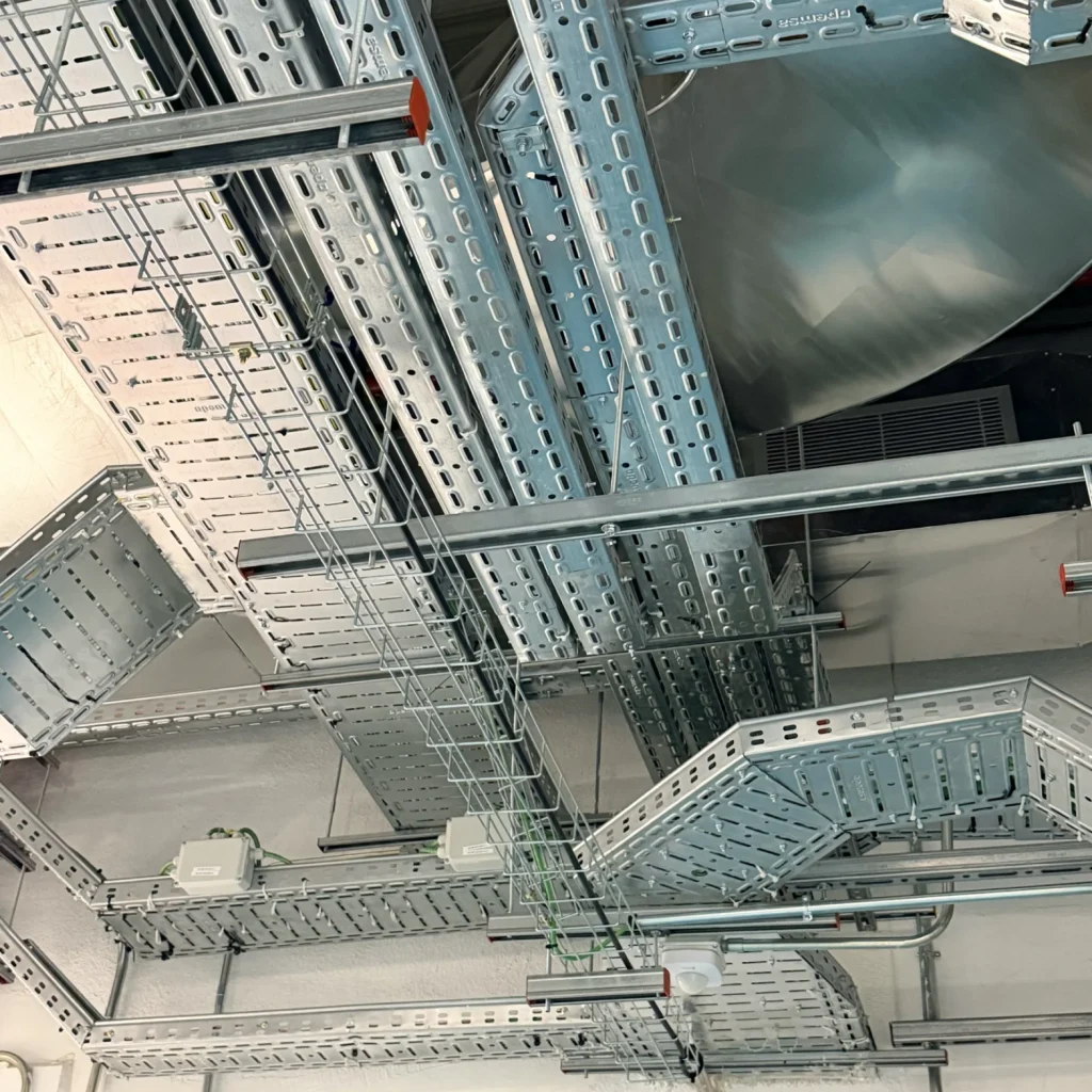 Intrincado montaje de bandejas portacables de metal sólido y de malla en el techo, con conductos de ventilación grandes. Se observan cajas de empalme blancas y el logo de "TORRECILLA INSTALACIONES ELÉCTRICAS".