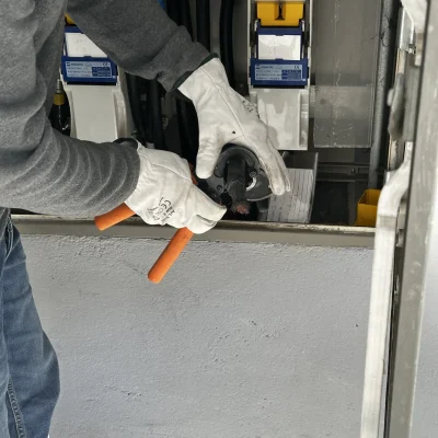 Un técnico con guantes de seguridad blancos sostiene una herramienta pelacables junto a un cuadro eléctrico abierto.
